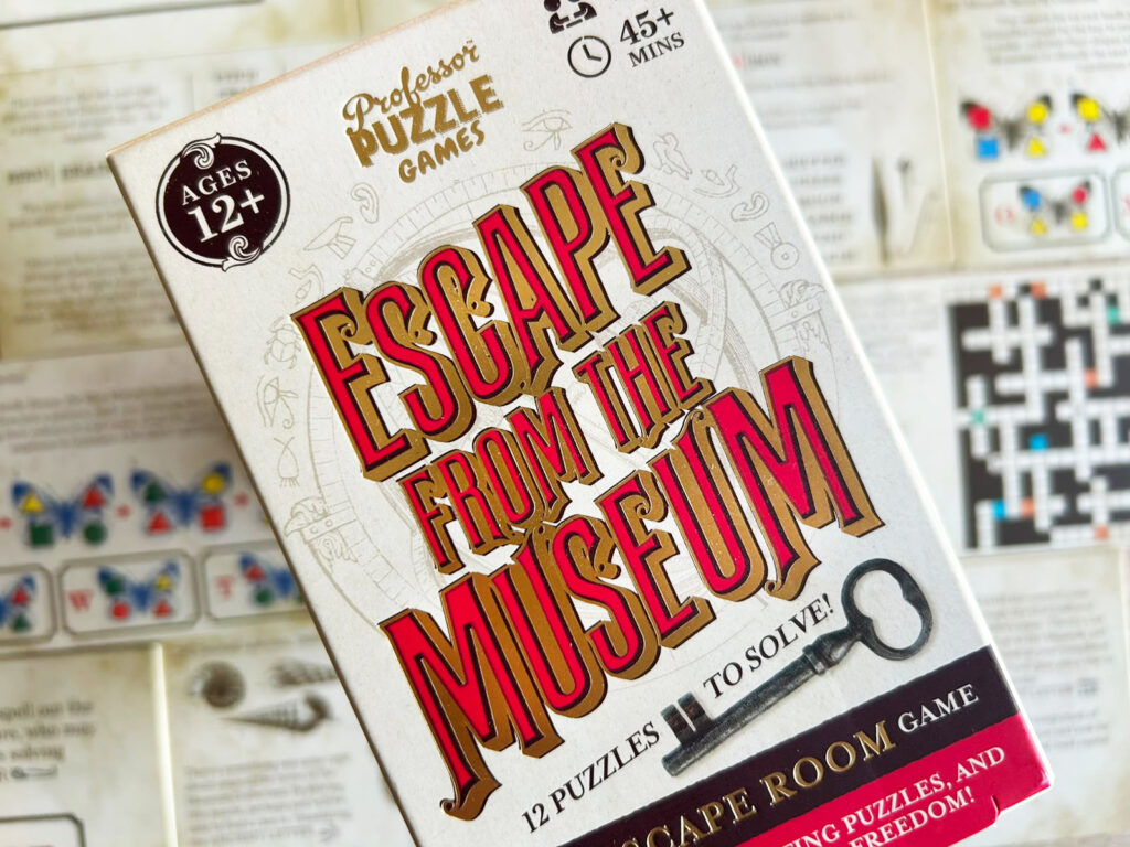 Die Verpackung des Spiels „Escape from the Museum“ von Professor Puzzle, im Hintergrund sind die Rätselkarten zu sehen.