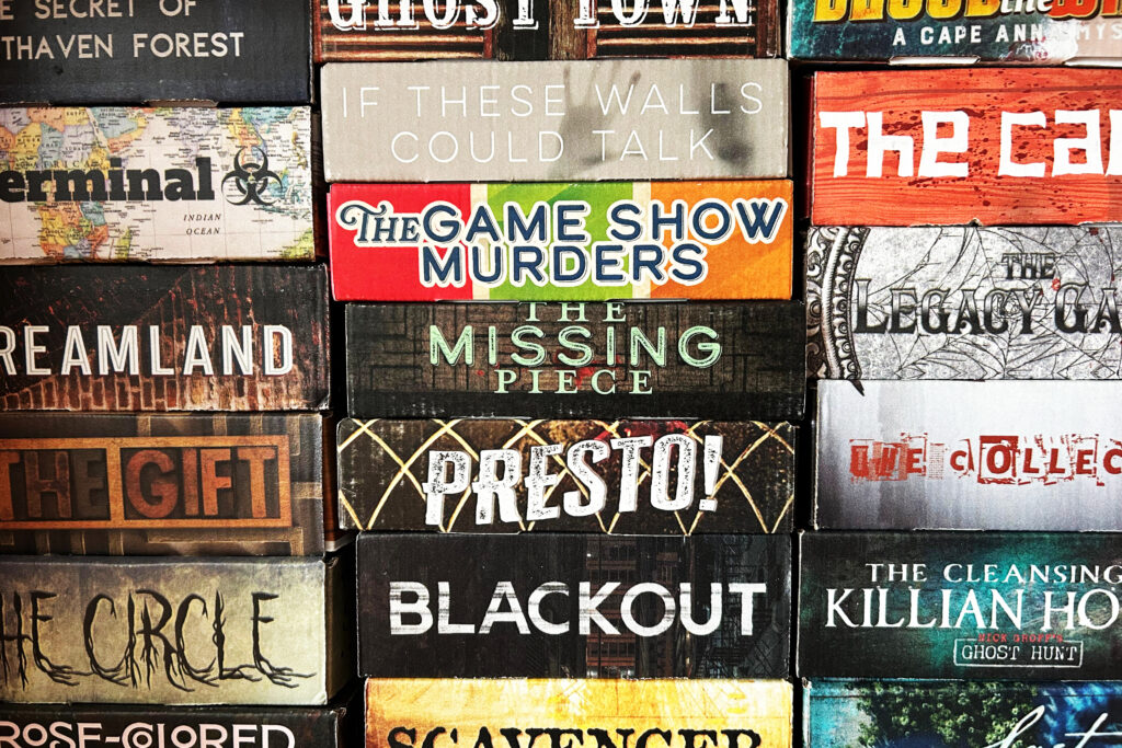 Das Foto zeigt einen Ausschnitt einer Spielesammlung: In einem Regal stapeln sich verschiedene bunte Schachteln des Anbieters Deadbolt Mystery Society.