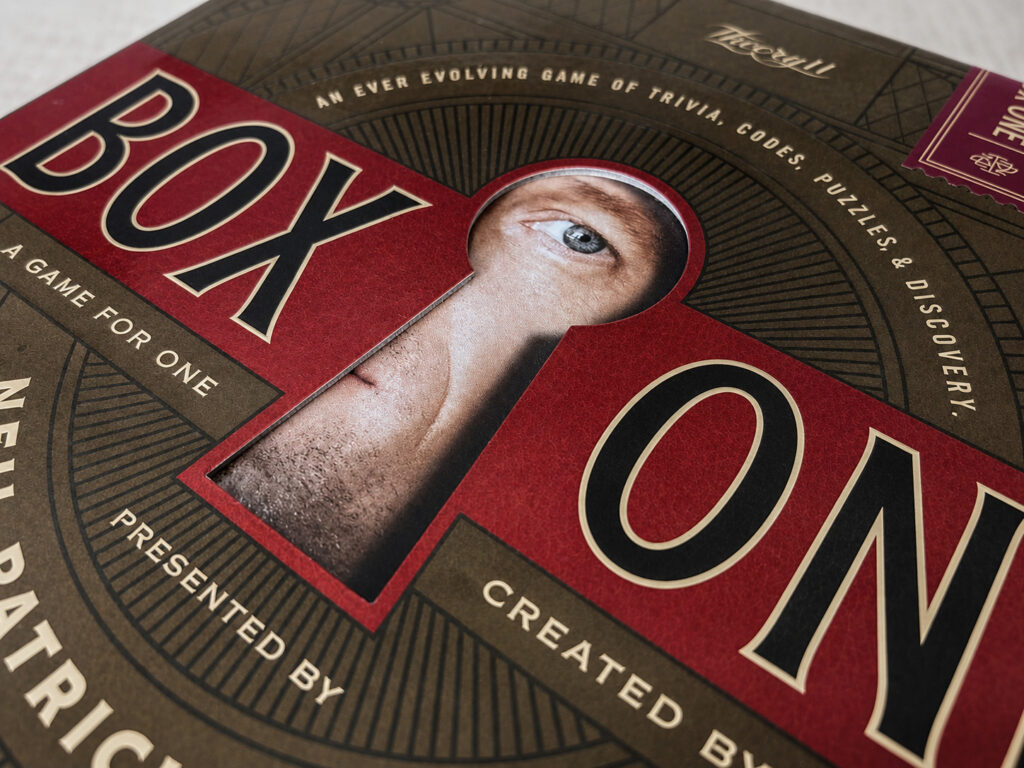Das Cover der Spielebox von „Box One“ von Neil Patrick Harris. Der Schauspieler scheint durch ein Schlüsselloch im Deckel der Schachtel zu schauen.