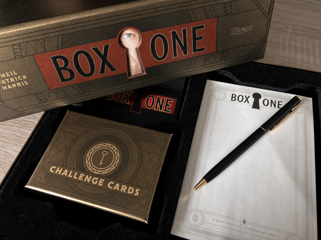 Ein Blick in die geöffnete Spielebox von „Box One“. Zu sehen sind eine kleine Schachtel mit Karten, ein schwarzer Umschlag, ein Notizblock und ein Stift.