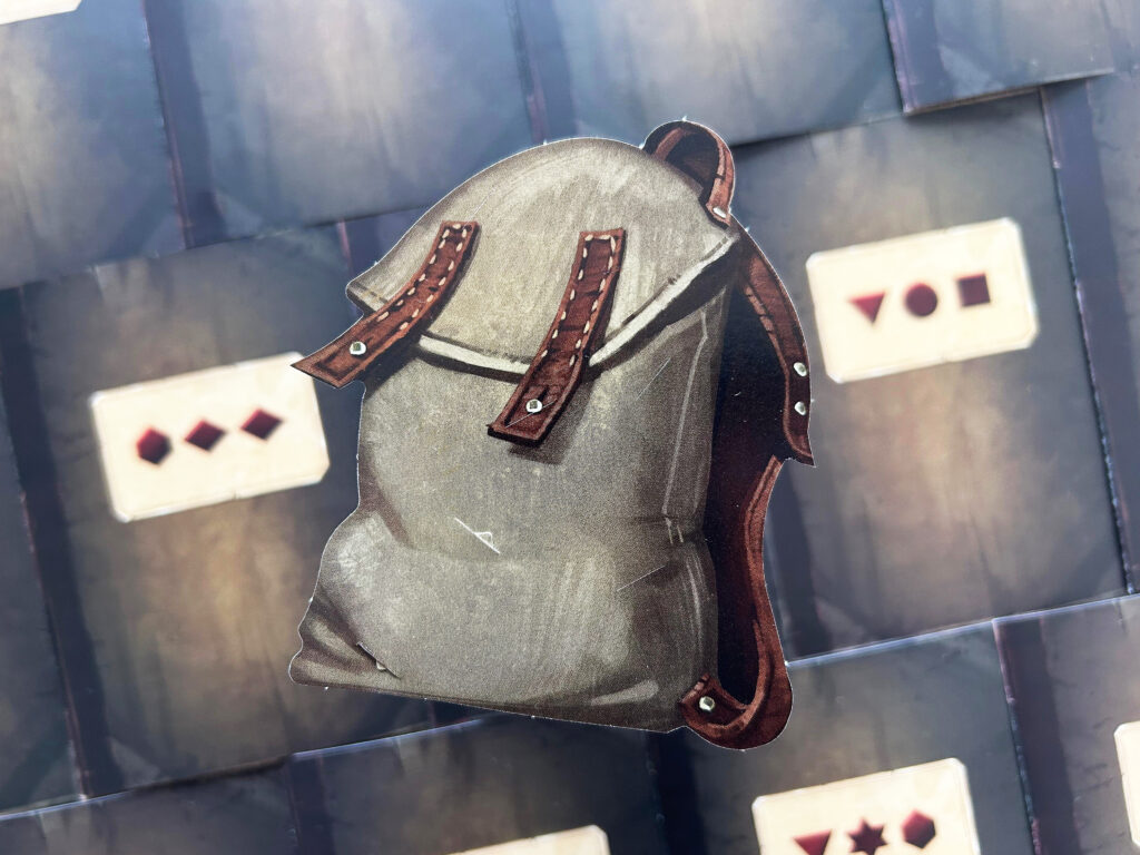 Ein kleiner Rucksack aus Pappe liegt auf dem ausgebreiteten Spielmaterial von „Das dunkle Schloss“ von EXIT.