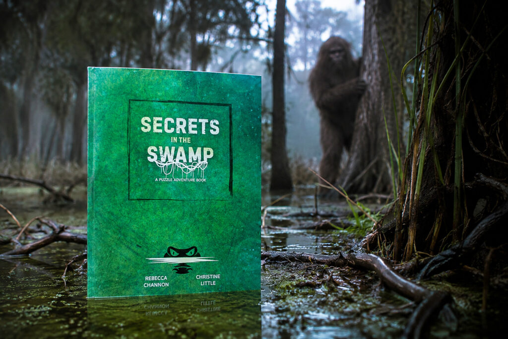 Das Buch „Secrets in the Swamp“ ist in einer Sumpflandschaft zu sehen, im Hintergrund schaut eine haarige Figur hinter einem Baum hervor.