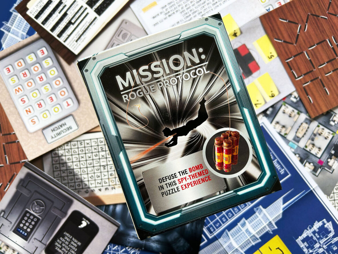 Die Schachtel des Spiels „Mission: Rogue Protocol“ von Red Herring Games liegt auf dem ausgebreiteten Spielmaterial.