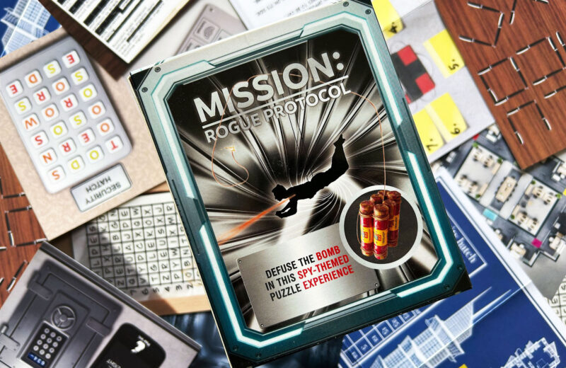 Die Schachtel des Spiels „Mission: Rogue Protocol“ von Red Herring Games liegt auf dem ausgebreiteten Spielmaterial.